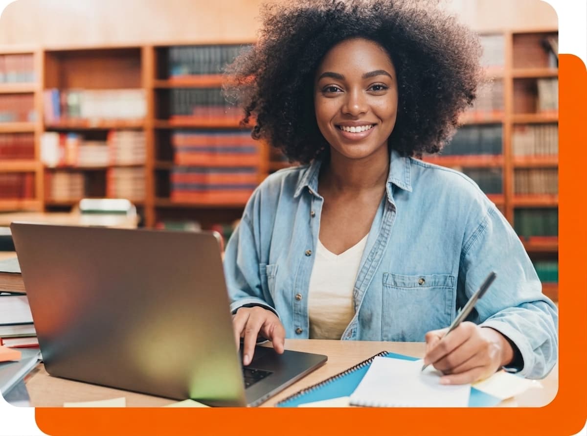 Estudante sorrindo em uma biblioteca, com um notebook aberto sobre a mesa e escrevendo em um caderno