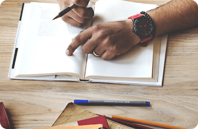 Pessoa estudando com um livro aberto, lápis na mão e canetas sobre a mesa